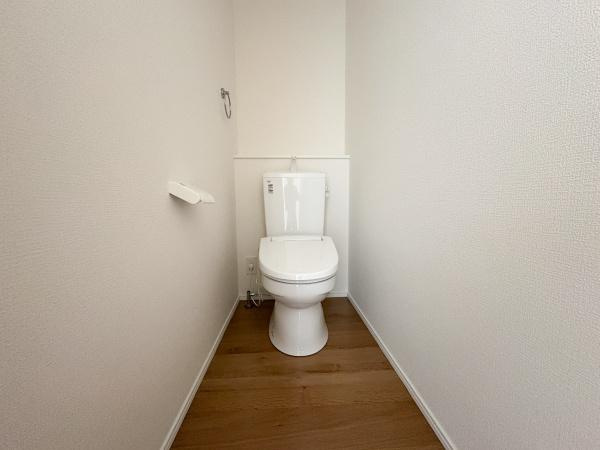 大字鴛野1期2号棟のトイレ|トイレはホワイトを基調とした清潔感のある空間です。床材の木目調がやさしい雰囲気を演出します。シンプルなデザインで掃除がしやすい造りです。ペーパーホルダーやタオル掛けも設置されています。