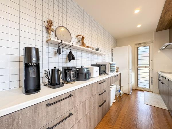 角子原　戸建てのキッチン|白を基調としたタイル壁と木目の収納が調和したキッチンです。広々とした作業スペースが確保され家電の配置もしやすい設計です。明るい照明と清潔感のある空間が印象的です。収納が豊富で整理整頓がしやすいです。