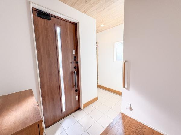 角子原　戸建ての玄関|玄関ドアの木目調が印象的で空間全体に明るさがあります。白を基調とした壁とタイルが清潔感を感じさせます。段差部分には手すりが設置されていて安心です。収納スペースも備わっていて整理整頓しやすい設計です。