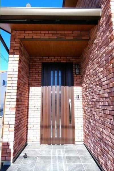 HT久原4号地の玄関|レンガ調の外壁が印象的な玄関です。シンプルなデザインの扉が建物全体に調和しています。屋根付きなので天候にも配慮されています。玄関横に設置された照明が夜間の出入りもサポートします。