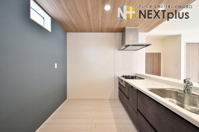 白岡市小久喜4期のキッチン|木目調の下がり天井がカフェのようなお洒落な空間を演出。グレーのアクセントクロスが空間を引き締め、洗練された印象を与えます。勝手口や小窓から光が入り、明るく開放的な気分でお料理を楽しめるキッチンです。