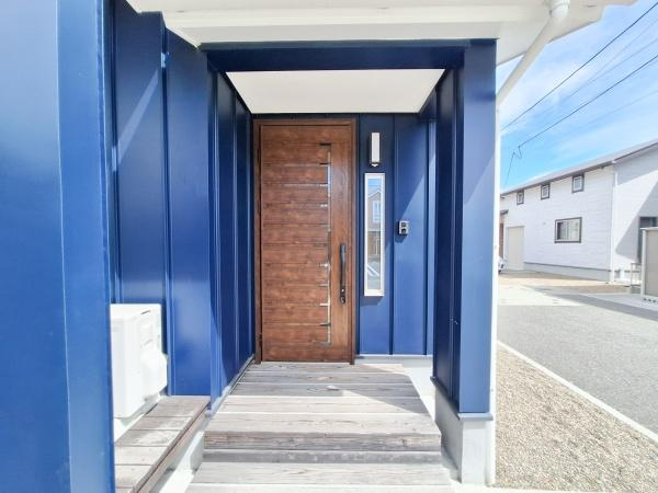角子原二丁目　戸建ての玄関|ダークトーンの外壁と木目調のドアが印象的な玄関です。広めのアプローチで出入りがスムーズに行えます。照明やインターホンが設置され防犯面にも配慮されています。