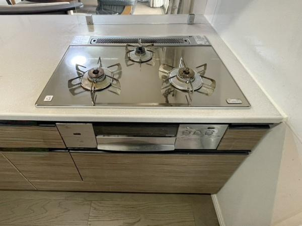 グリーンヒル大分駅前レジデンスのキッチン|三口コンロが備わったキッチンです。調理スペースも広々としていて効率的に作業が進められます。ガスコンロまわりはフラットで拭き掃除も簡単です。木目調の収納もあり見た目がすっきりしています。