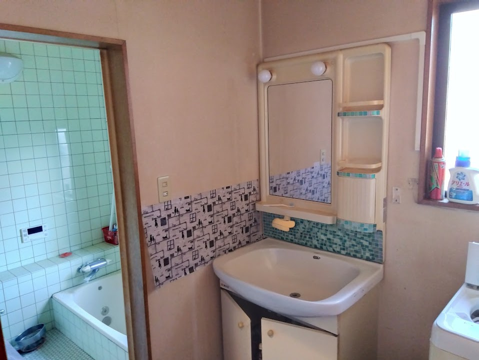 売中古戸建　三島市若松町の洗面所|朝の身支度も気持ちよく行える、清潔感のある空間です。収納棚付きの洗面台で、日用品やタオル類もすっきり整理可能ですね。アクセントクロスとタイルが空間のポイントとなり、程よいレトロ感を演出しています。