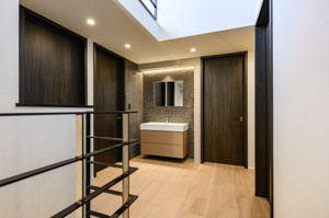 【洗面所】 | 黒のアイアン手すりがアクセントとなり、空間を引き締めます。吹き抜け越しに上下階の繋がりを感じられる設計で、ご家族の気配を程よく共有。木の質感が心地よい廊下は、歩くたびに住まいの品質を実感できます。