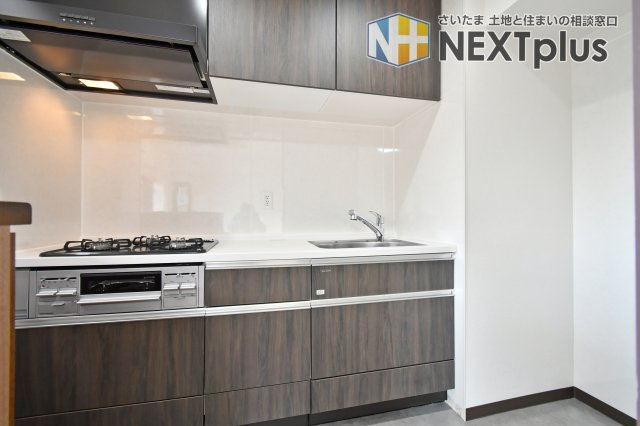 光建ハイムブリリアンス大宮公園のキッチン|シックな木目調パネルが空間を引き締める、2025年2月に刷新されたシステムキッチン。3口コンロ付きで効率良く調理でき、壁面は清掃しやすいパネル仕様。お料理が楽しくなる、機能性と美しさを兼ね備えた空間。