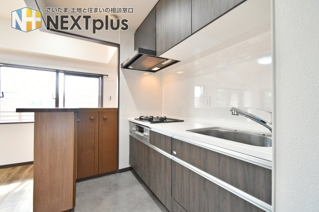 光建ハイムブリリアンス大宮公園のキッチン|シックな木目調パネルが洗練された空間を演出するシステムキッチン。三口コンロ採用で日々のお料理も効率よく楽しめます。リビングを見渡せるカウンター付きで、ご家族との会話を楽しみながら家事ができる設計です。