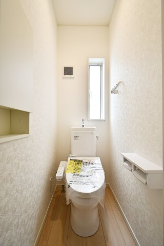 伊奈町本町第3　全2棟　1号棟のトイレ|高機能温水洗浄便座を標準装備した、白基調の清潔感ある空間です。便利な壁面収納棚や、明るい日差しが入る窓付き。毎日使う場所だからこそ、換気効率と心地よさにこだわった設計が魅力的なポイントです。