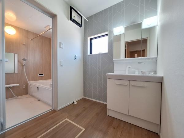 大字佐野1期3号棟の独立洗面台|洗面所は清潔感のある空間でシンプルなデザインが印象的です。窓からの採光があり明るい室内です。大型のミラーキャビネットで使いやすさが感じられます。床材の木目調も落ち着いた雰囲気を演出しています。