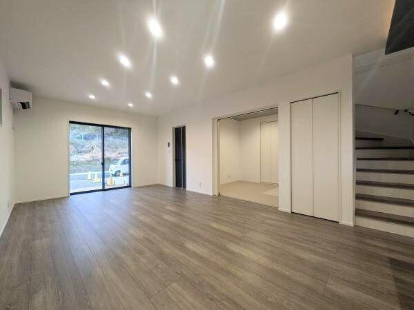 タマタウン横尾東町2　2号地の居間・リビング|明るい照明と大きな窓が特徴のリビングです。床は落ち着いた色合いのフローリング仕様となっています。リビング横には便利なスペースが設けられています。収納もあり機能的な空間です。階段への動線もスムーズです。