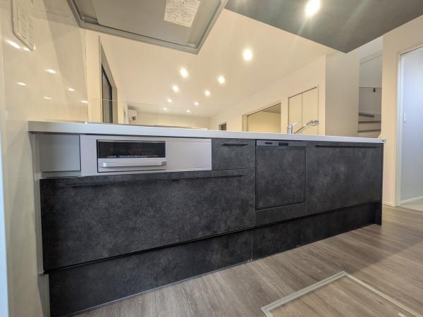 タマタウン横尾東町2　2号地のキッチン|落ち着いた色合いのキッチンが室内に調和しています。天井照明が印象的で全体を明るく照らします。キッチンカウンターは広くすっきりとした印象です。収納スペースも豊富に確保されています。