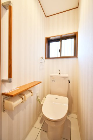 さいたま市見沼区深作2丁目のトイレ|温水洗浄便座へ一新された、清潔感あふれるトイレ。窓からの採光と換気も良好で、ストライプ柄のクロスが空間を明るく彩ります。手すり付きで幅広い世代に優しい設計です。