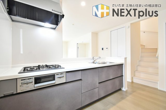 第17上尾上　全6棟　B号棟のキッチン|キッチン横に階段を配置した、家族のコミュニケーションを育む間取りです。三口コンロやワイドシンクなど充実の設備に加え、リビングまで真っ直ぐ見渡せる設計が、忙しい家事の時間にゆとりと安心感を与えます。