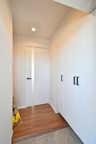 春日部ダイヤモンドマンションの玄関|木目調のフローリングと黒のドアハンドルがアクセントのモダンなデザイン。スリット入りのリビングドアから優しい光が漏れ、帰宅した瞬間に家族の温もりを感じられる設計です。