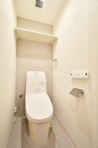 クラーク春日部壱番館のトイレ|温水洗浄便座一体型トイレへ新規交換。背面のアクセントクロスが空間に彩りを添え、上部の棚はストック品の収納に便利です。フロアタイルも貼替済みで、毎日使う場所だからこその清潔感と快適さを追求しました。