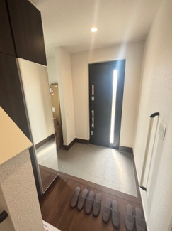 【玄関】 | 東広島市八本松飯田6丁目（全2区画）1号棟　 | 姿見付きの大型シューズボックスを備えた玄関。落ち着いた木目調のフローリングとホワイトの壁紙が、清潔感のある空間を演出します。手すりやダウンライトなど、細かな設備も整っています。