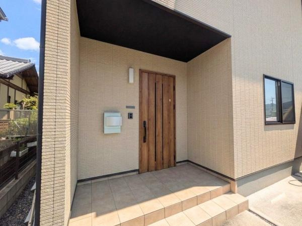 大字江無田　戸建ての玄関|木目調の玄関ドアが外観に調和しています。広々としたタイルのアプローチが特徴です。屋根付きのポーチで雨の日も安心して出入りできます。シンプルなデザインが周囲と調和しています。