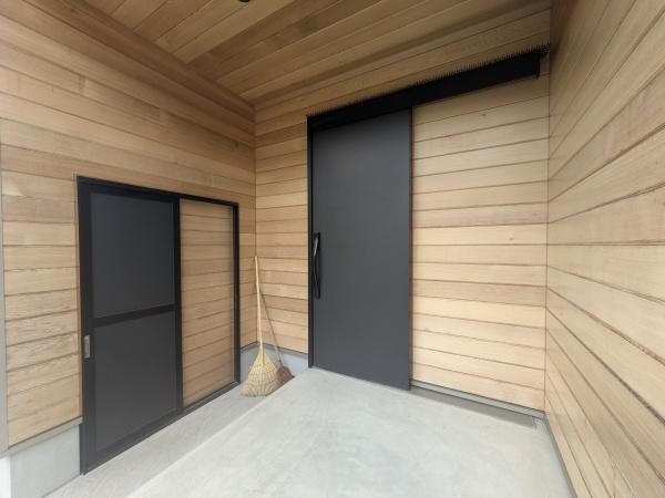 久原南2丁目　戸建て(美容室併設)の玄関|木材をふんだんに使用した外壁が玄関スペース全体に温かみを感じさせます。シンプルなデザインの玄関ドアは落ち着きある雰囲気を醸し出しています。床はフラットで使いやすく幅広い世代に配慮されています。