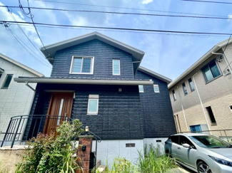 【外観】 | あすみが丘東　リフォーム済み【3LDK】中古戸建：JR外房線「土気」駅 | 黒を基調とした凹凸のある外壁と、温かみのある木目調の玄関ドアが対照的な2階建て住宅の外観です。アプローチには石畳が敷かれ、緩やかにカーブする黒いアイアン手すりが特徴的です。