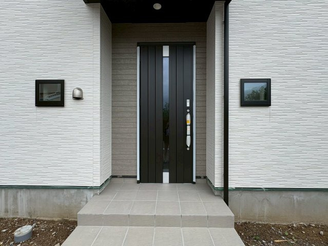 「３帖の畳コーナー付」高知市東城山町3号棟　新築一戸建ての玄関|木目調のアクセント壁と黒いドアが映える、モダンな外観デザイン。