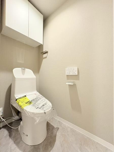 綱島ハイムのトイレ|清潔感のあるトイレ空間です。収納棚が備え付けられており整理しやすい仕様になっています。壁付けの操作パネルで操作もしやすいです。スペースにゆとりがありすっきりとした印象です。