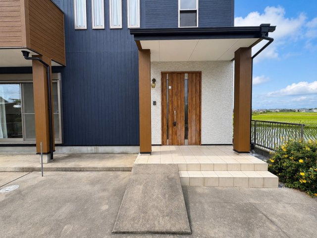 吉川市　二ツ沼　中古戸建のエントランス|【エントランスアプローチ】木目の玄関ドアとネイビーの外壁が印象的なエントランス。屋根付きのため雨の日でも出入りしやすく、玄関前にはゆとりあるスペースを確保。