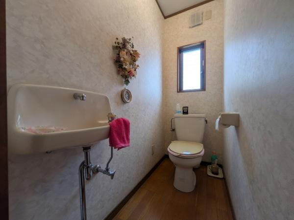 皆春　戸建てのトイレ|窓から自然光が差し込む明るいトイレです。壁に飾られた花の装飾が空間を彩ります。手洗い場も併設されていて便利です。床材は木目調で温かみがあります。清潔感のある内装です。