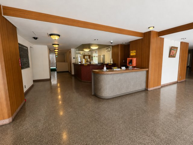 虹ヶ丘マンション本館のエントランス|管理員室と受付カウンターを備えた共用ロビーです。木目調の内装が落ち着いた雰囲気を演出しており、AEDも設置されています。