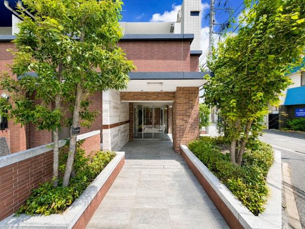 レ・ジェイド横濱花之木のエントランス|植栽の緑と重厚感あるレンガ調の外壁が迎えるエントランスアプローチ。落ち着いた雰囲気と清潔感があり、住まう方や訪れる方を上品に迎えてくれる、マンションの第一印象を高める空間です。