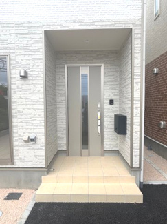【玄関】 | 大竹市小方1丁目【全4区画】No4　 | 屋根付きのインナーアプローチ仕様の玄関。雨の日でも濡れにくく、落ち着いて鍵の開け閉めができます。外壁の凹凸感ある意匠とシックな色調のドアが、上品なエントランスを演出します。