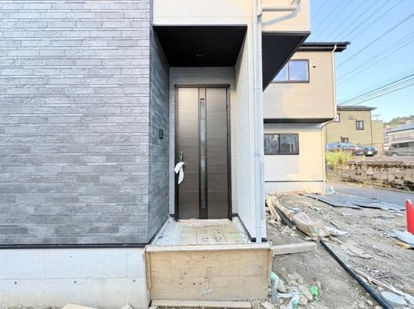 横浜市金沢区釜利谷南２丁目の新築一戸建の玄関|スタイリッシュなデザインの玄関ドアが設置されています。外壁と調和した色合いが特徴的です。玄関周りはシンプルな造りですっきりとした印象です。周囲のスペースも広めに確保されています。