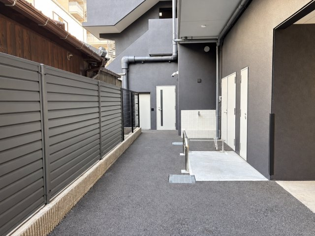 ワコーレ阿倍野播摩町ラフィリスのその他|ダークグレーの外壁と目隠しフェンスが印象的な共用スペースです。建物のデザインに統一感があり、落ち着いた雰囲気に仕上がっています。屋外に設けられた設備や通路の配置も確認できます。