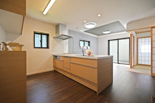 尼崎市東園田町2丁目新築一戸建て│4LDK+駐車2台（車種制限有）のキッチン|1階LDKをキッチンユニット越しに撮影した内観写真です。フルフラットの対面式オープンキッチンを採用しているため視界は広く、ご家族間やご来訪者とのコミュニケーションも一体的に取り易くなっております。