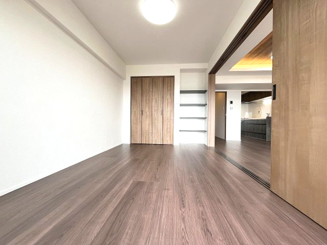 忠道マンションA棟の洋室|落ち着いた色合いのフローリングが広がる明るい洋室です。クローゼットとオープンシェルフが備わり、収納も充実。隣接するリビングとの一体感も感じられます。