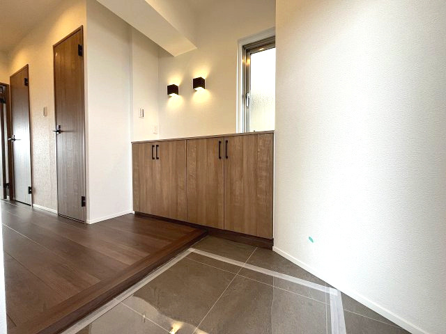 忠道マンションA棟の玄関|木目の美しいシューズボックスと壁を照らすブラケットライトがモダンな印象を与える玄関です。床はタイルとフローリングのコントラストが美しく空間に奥行きを感じさせます。窓があるので自然光も取り入れられます。