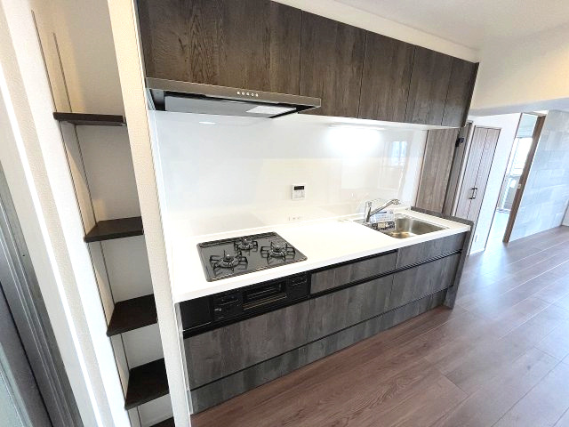 忠道マンションA棟のキッチン|落ち着いた木目調のキャビネットと清潔感のある白いワークトップが印象的なキッチンです。使い勝手の良い3口コンロや、十分な収納スペースが料理の時間を快適にします。