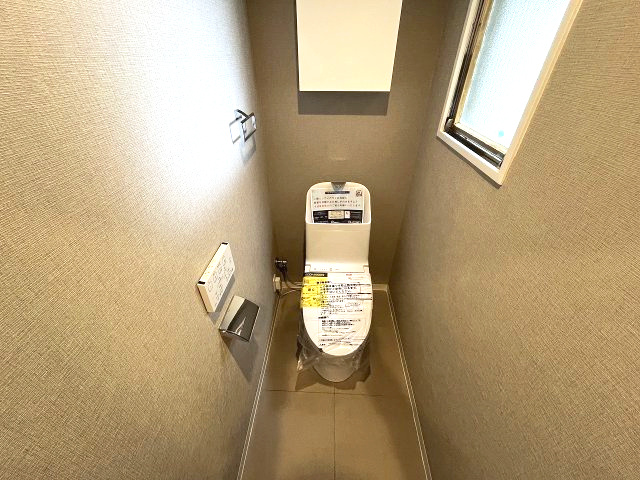 忠道マンションA棟のトイレ|壁一面に施された落ち着いたデザインのクロスが、落ち着きと上質さを感じさせるトイレ空間。最新の温水洗浄便座が備わり、快適性も兼ね備えています。窓があるため明るく開放的な印象です。