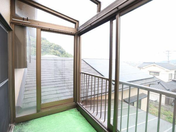 葛木　中古のバルコニー|広々としたバルコニーは採光性に優れ明るい空間が広がります。屋根付きで天候の影響も受けにくく活用しやすいです。ガラス張りの柵で開放感があり外の景色を楽しめます。グリーンの床材がアクセントとなっています。