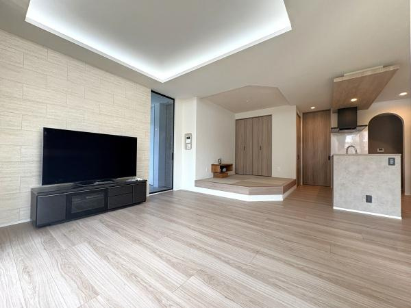 寒田南町二丁目　中古の居間・リビング|木目調のフローリングと間接照明が落ち着いた雰囲気を作り、大きなテレビとシンプルなテレビ台が配置されたリビング。和室風の段差でくつろげ、オープンなレイアウトが広がりを感じさせます♪