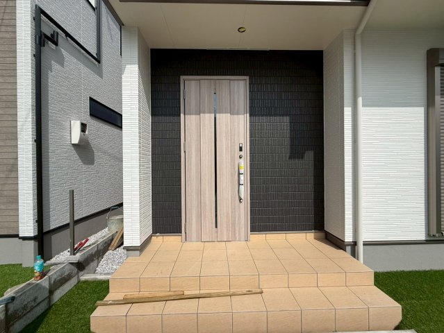 香美市土佐山田町宝町2期5棟 2号棟の玄関|木目調の玄関ドアとブラック外壁が調和した、上品なデザインの玄関。屋根付きポーチで雨の日も濡れにくく、ゆとりのある階段設計。