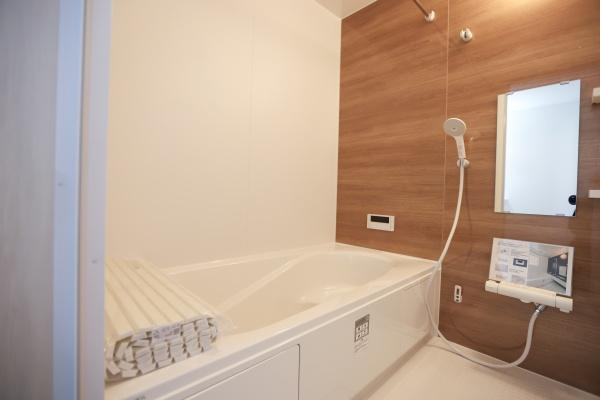 大字森町4期1号棟の浴室|木目調のアクセントパネルが落ち着いた雰囲気を演出しています。浴室乾燥機が備え付けられており機能的です。使いやすいワイドな浴槽で清潔感のある空間です。便利なミラーと収納棚も設置されています。