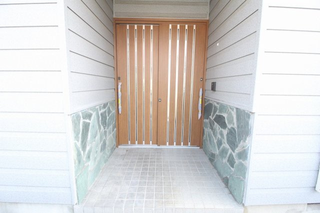 小山市中久喜1丁目の玄関|玄関ドアは木目調デザインで温かみのある印象。庇付きの造りで、雨の日の出入りも安心です。落ち着いた色合いが外観にしっくりと馴染んでいます。