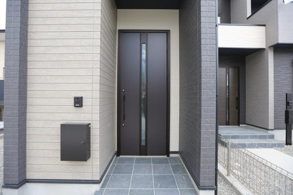 横尾東町2丁目2期1号棟の玄関|モダンな外観が印象的な玄関です。ダークトーンのドアが全体を引き締めています。タイル張りのアプローチが清潔感を感じさせます。宅配ボックスも設置されています。雨除けスペースも確保されています。
