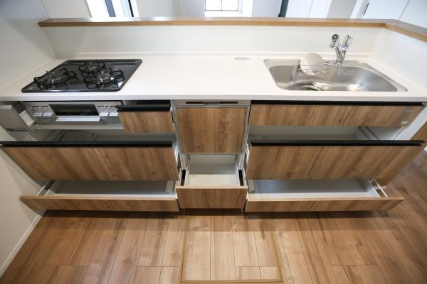 横尾東町2丁目2期3号棟|木目調のデザインがあたたかみを感じさせるキッチンです。大型の引き出しが複数あり収納スペースが充実しています。シンクとコンロが横並びでまとまっており作業効率をサポートします。