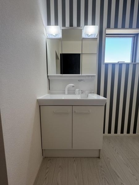 大字小池原1期3号棟の独立洗面台|ストライプ柄の壁紙がアクセントとなった洗面所は清潔感のあるホワイトの洗面台が調和しています。ミラー上には照明が2つ設置されており明るさもしっかり確保されています。