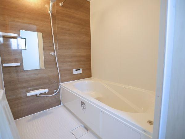 大字光吉4期2号棟の浴室|木目調のアクセントパネルが空間を引き締めます。広々とした浴槽とシャワーが快適なバスタイムをサポートします。明るく清潔感のあるデザインで日々のお手入れも簡単です。便利な鏡と棚も備わっています。