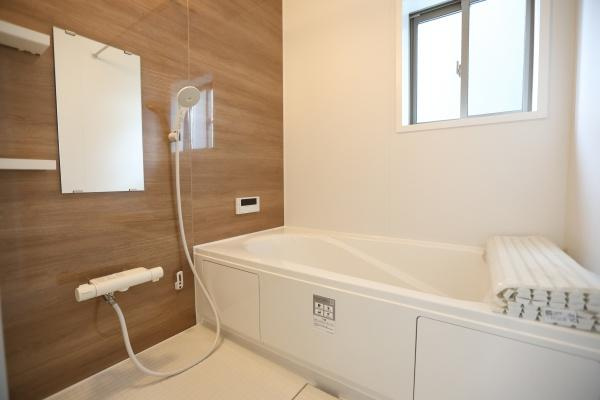 徳島2丁目1期2号棟の浴室|木目調のアクセントパネルが印象的なバスルームです。浴槽はゆったりとしたサイズで窓から自然光が入ります。シンプルで清潔感のある空間です。鏡や棚も設置されていて使い勝手にも配慮されています。