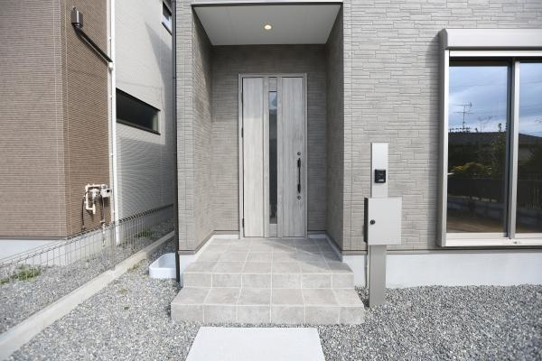 徳島2丁目1期3号棟の玄関|落ち着いた色合いの玄関が印象的です。雨の日も安心の屋根付きで実用的なデザインが魅力です。広めのタイルスペースが使い勝手の良さを感じさせます。スタイリッシュな外観が印象に残ります。