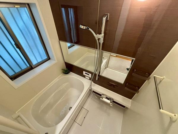 【浴室】 | 大きな鏡が付いたバスルームで開放感があります。落ち着いた色合いの壁面が特徴的です。浴槽はゆったりとしたサイズ感となっています。窓もあり換気がしやすい設計です。収納棚も備わっています。