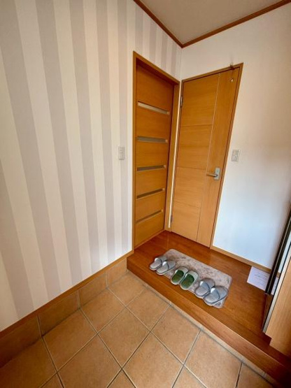 【玄関】 | 落ち着いた木目調のドアとストライプの壁紙が玄関スペースに明るさを演出しています。タイル張りの床はお手入れがしやすく清潔感があります。段差部分にスリッパを整然と並べることができ実用的な設計です。